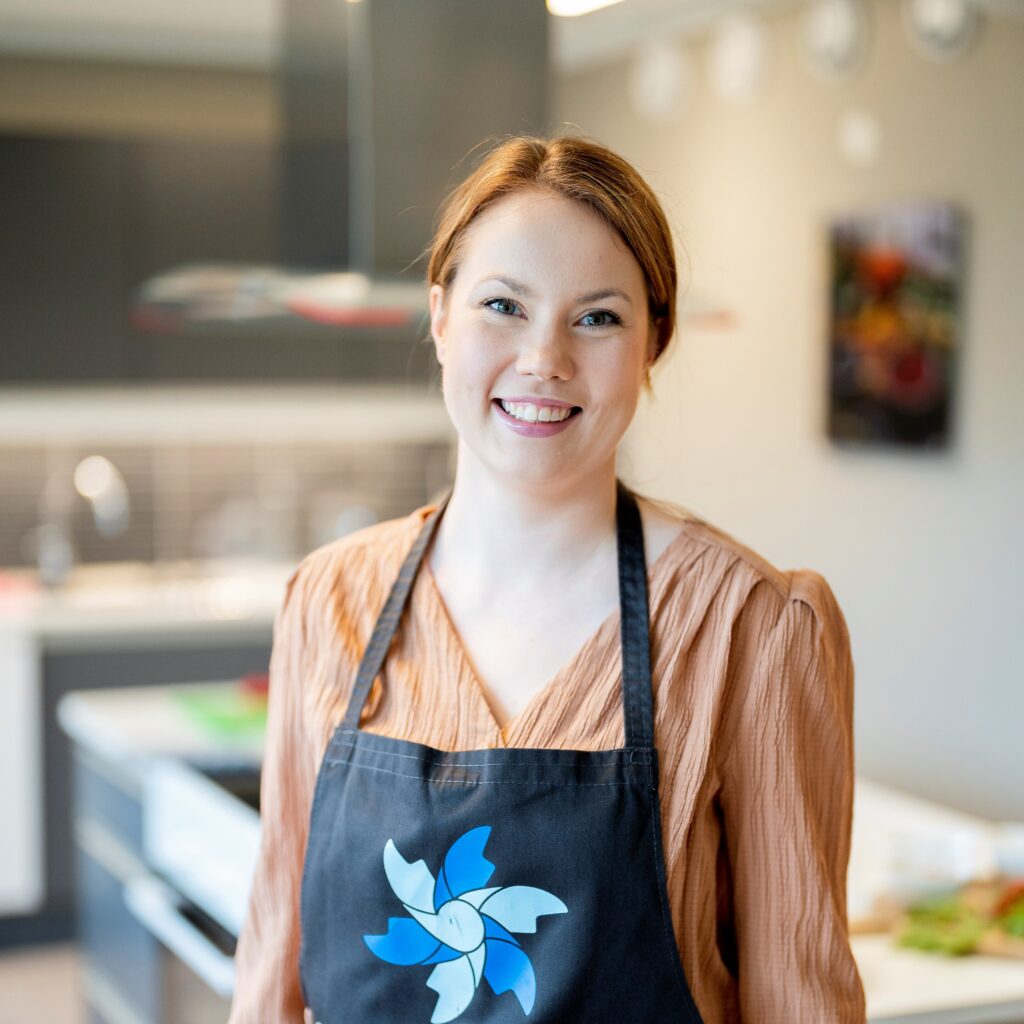 En kvinne i brun bluse og svart forkle står smilende i et moderne kjøkken.