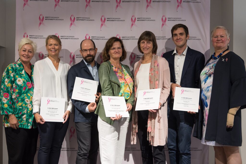 Sju personer står på rekke og rad med diplomer i hendene og poserer for et gruppebilde foran et bakteppe med logoer fra Brystkreftforeningens Rosa sløyfe.