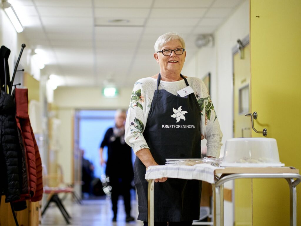 En eldre kvinne med briller og "Kreftforeningen"-forkle står i en gang ved siden av en vogn med tildekkede matvarer.