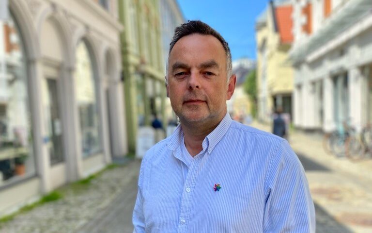 En mann i lyseblå skjorte står på en brosteinsbelagt gate med mange bygninger en solfylt dag.