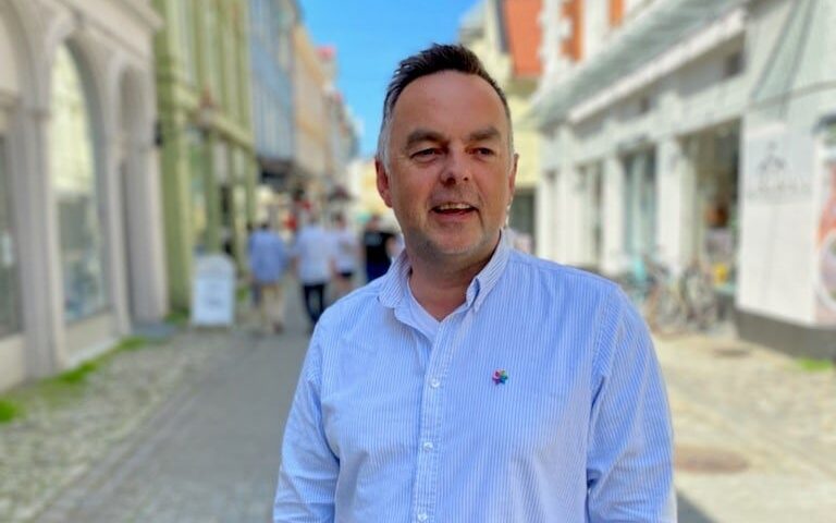 En mann i lyseblå skjorte står på en brosteinsbelagt gate med uskarpe bygninger og fotgjengere i bakgrunnen.