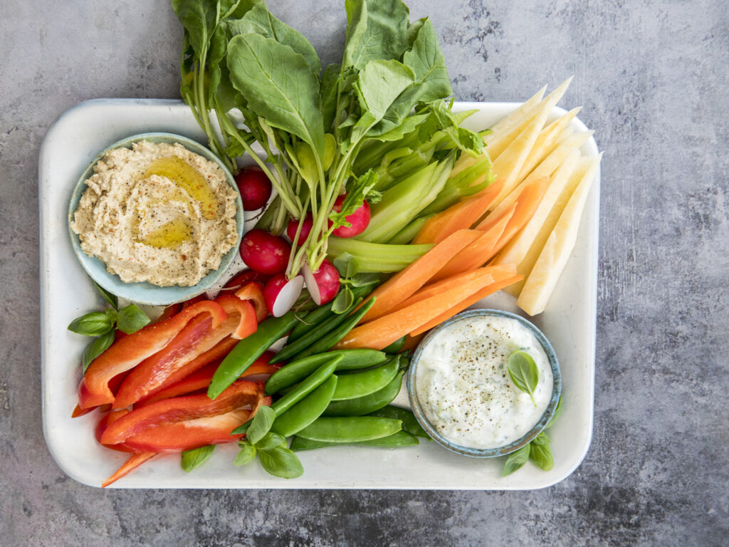 Et brett med et utvalg ferske grønnsaker, blant annet gulrøtter, selleri, reddiker, rød paprika og sukkererter, servert med boller med hummus og kremet dip.