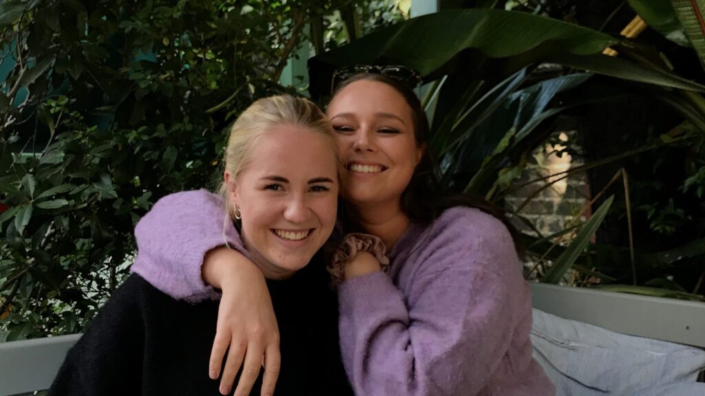 To kvinner sitter tett sammen på en benk utendørs og smiler til kameraet med den ene kvinnens arm rundt den andres skuldre. I bakgrunnen er det frodige, grønne planter.