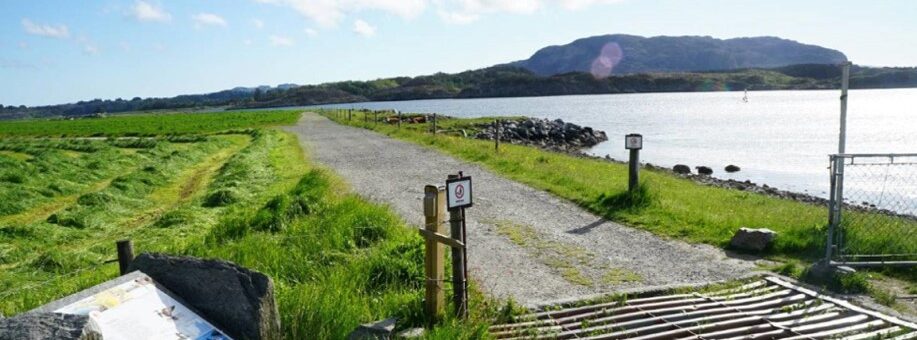 En grussti fører gjennom gressletter mot et vann med åser i bakgrunnen, og i forgrunnen ser man informasjonsskilt og et kveggitter.