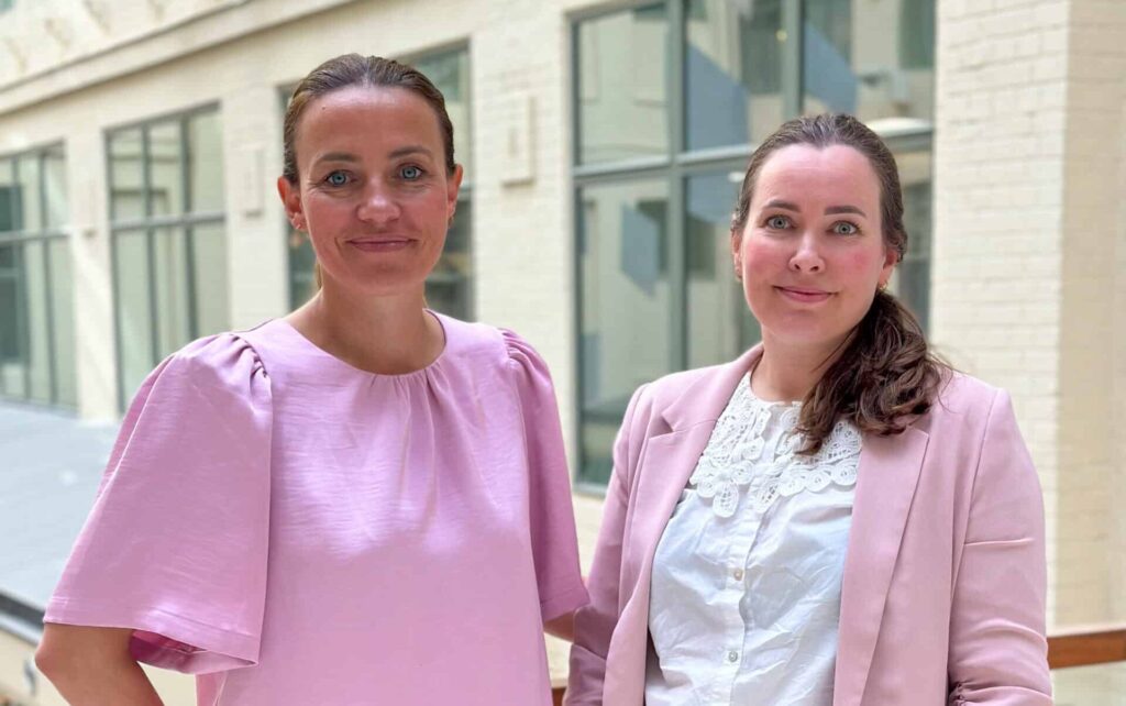 To kvinner står innendørs ved en vindusvegg, begge iført lyserosa overdeler og ser inn i kameraet.