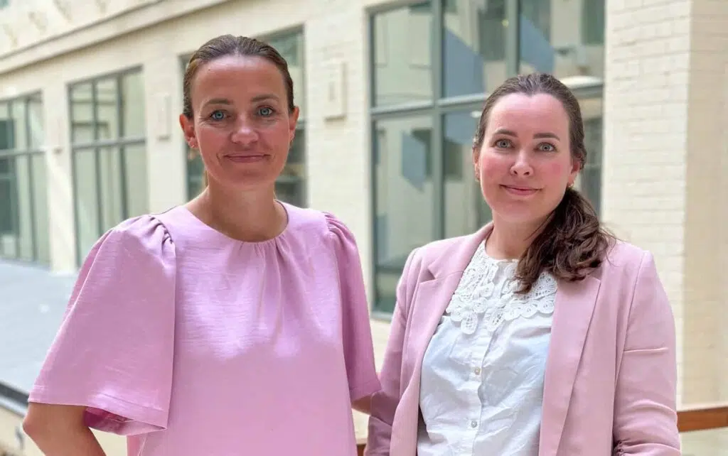 To kvinner står innendørs ved en vindusvegg, begge iført lyserosa overdeler og ser inn i kameraet.