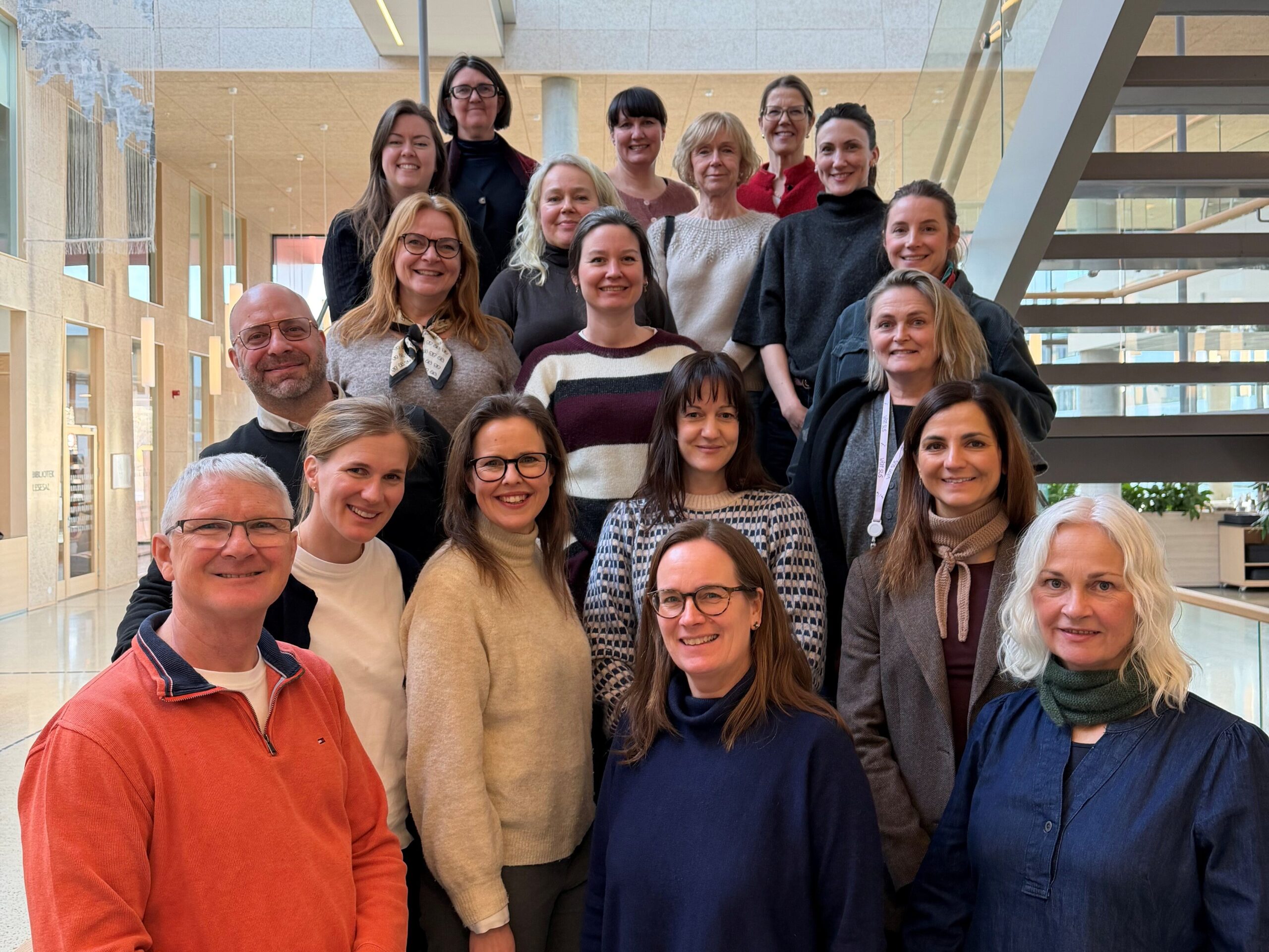 En gruppe på tjue personer står og smiler på en trapp inne i en moderne bygning med glassvegger og naturlig lys.