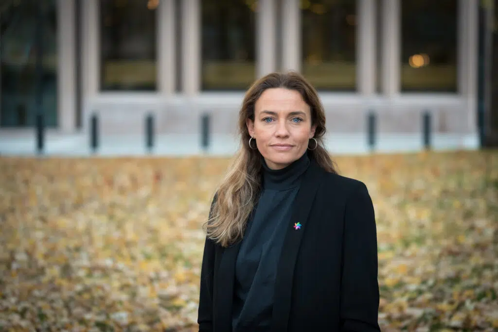 En kvinne med langt, brunt hår og svart blazer står ute på en gressplen dekket av høstløv, med en bygning i bakgrunnen.