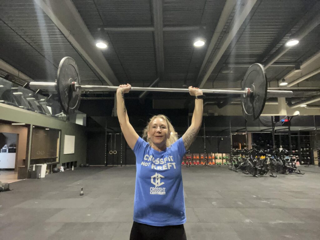 En person iført en blå "CrossFit not Kreft"-skjorte løfter en vektstang over hodet i et treningsstudio med treningsutstyr i bakgrunnen.
