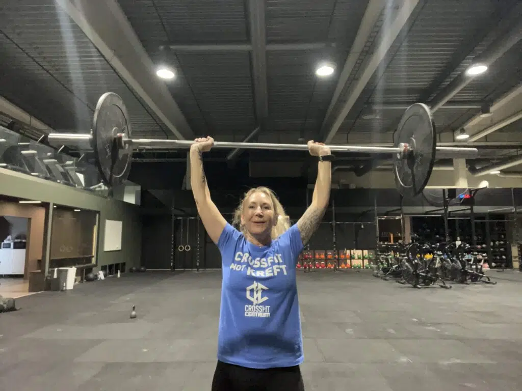 En person iført en blå "CrossFit not Kreft"-skjorte løfter en vektstang over hodet i et treningsstudio med treningsutstyr i bakgrunnen.