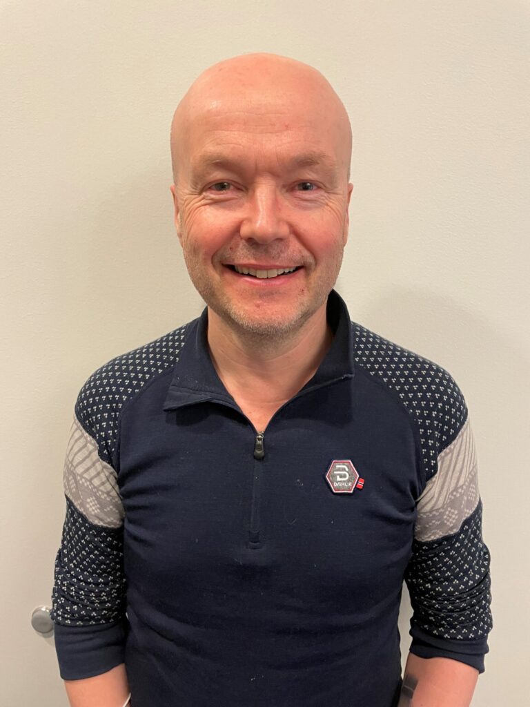 En skallet mann iført marineblå og grå langermet skjorte med en liten lapp på brystet står smilende foran en ensfarget, lys vegg.