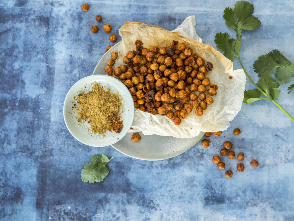 På en tallerken med bakepapir ligger ristede kikerter ved siden av en liten bolle med malte krydder, med friske korianderblader på en blå strukturert overflate.
