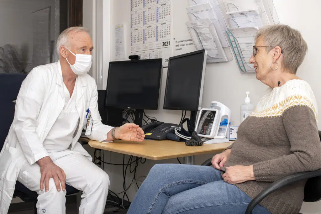 En lege i hvit frakk og maske snakker med en eldre kvinne som sitter på et kontor med dataskjermer og medisinsk utstyr på skrivebordet.