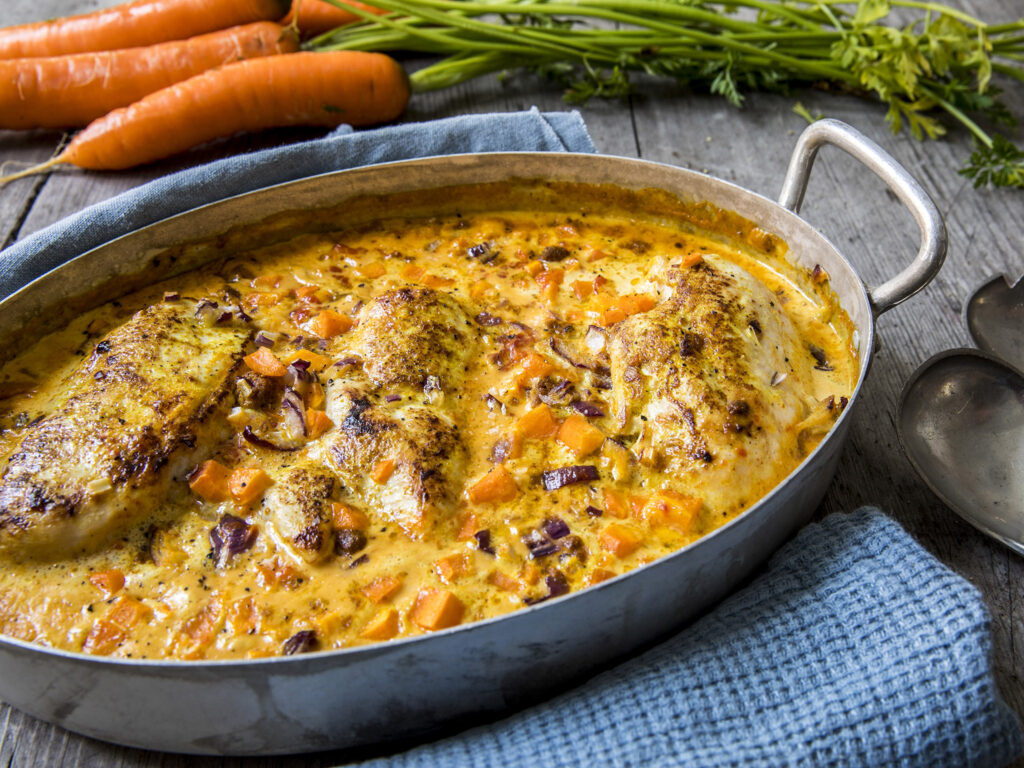 Bakte kyllingbryst i en kremet saus med terninger av gulrøtter og løk i et ovalt metallfat, med ferske gulrøtter og et blått håndkle ved siden av.