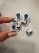 En hånd holder en liten hvit kopp med blått lokk og fargerik logo. Flere identiske miniatyrkopper står spredt på en hvit flate.