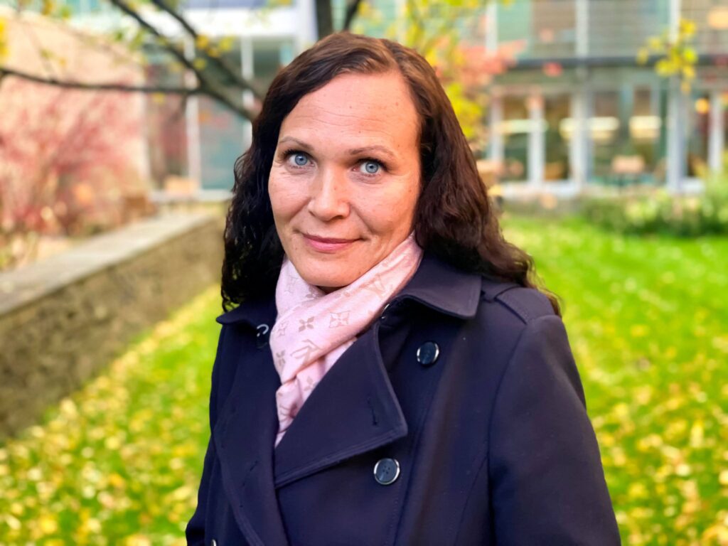 En kvinne med langt, mørkt hår, iført marineblå frakk og rosa skjerf, står ute på en gresslette med høstløv og bygninger i bakgrunnen.