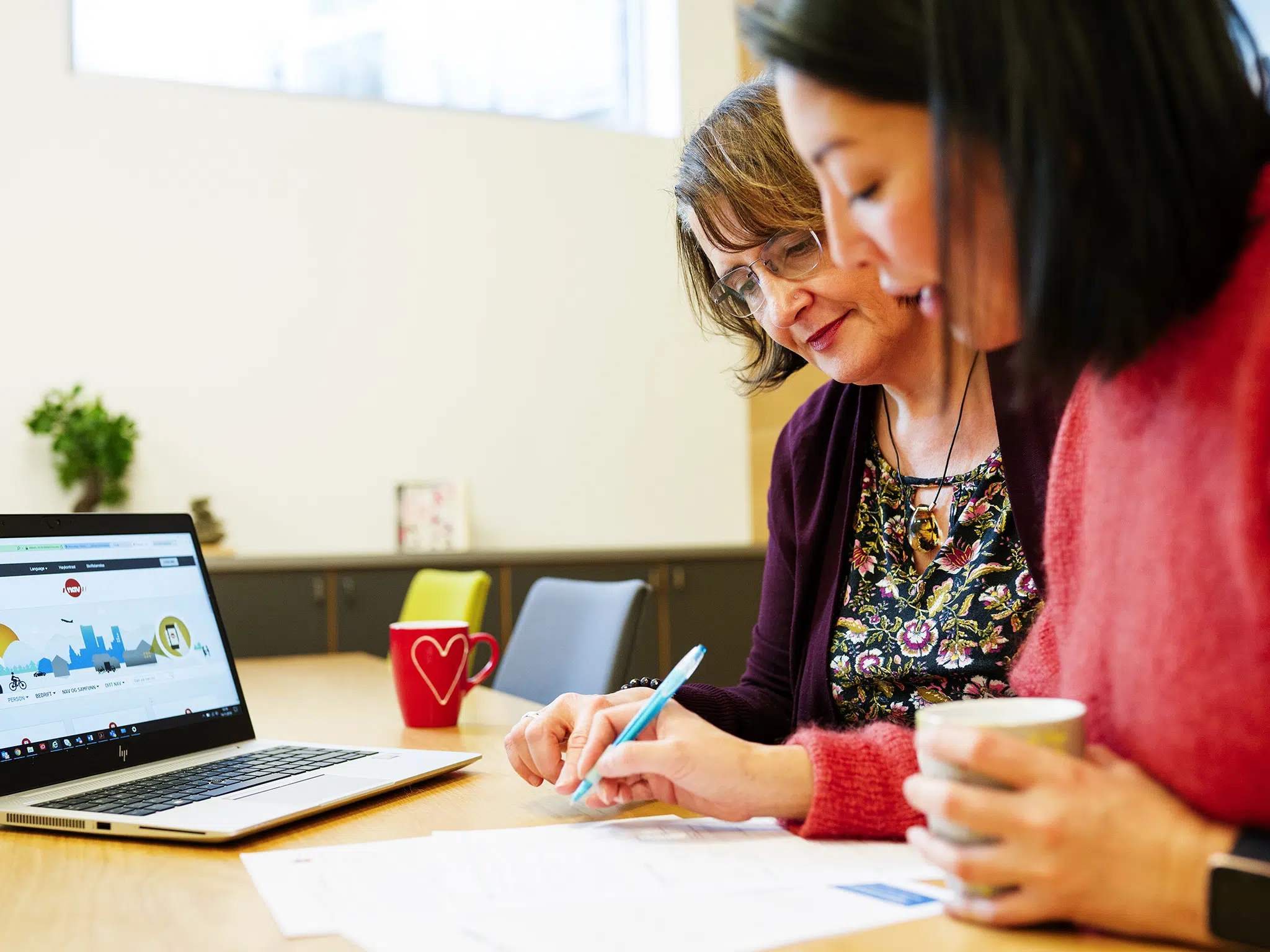 To kvinner sitter ved et bord og går gjennom dokumenter sammen, med en bærbar datamaskin og krus på bordet i et lyst kontormiljø.