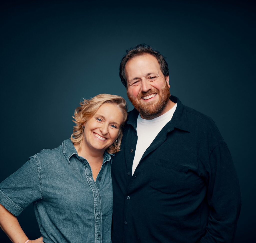 To voksne, en kvinne og en mann, står tett sammen og smiler til kameraet mot en mørk, ensfarget bakgrunn.