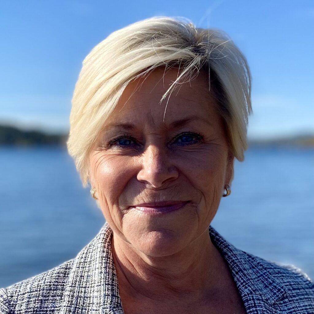 En kvinne med kort, blondt hår og rutete blazer smiler utendørs, med vann og blå himmel i bakgrunnen.