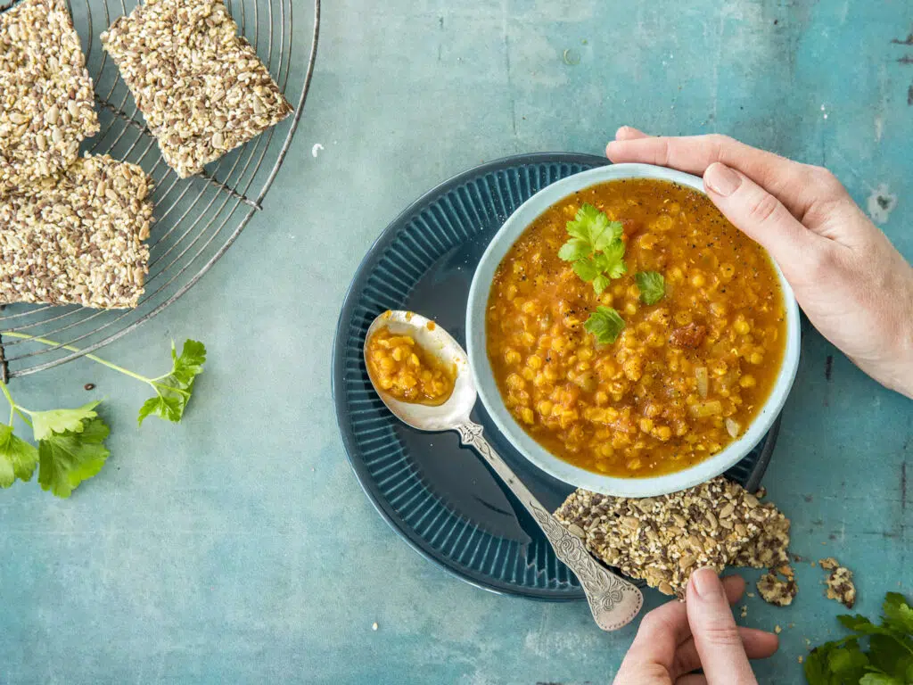 En person holder en kjeks med frø og en skål med oransje linsesuppe pyntet med persille, med flere kjeks og persille på en blå flate.