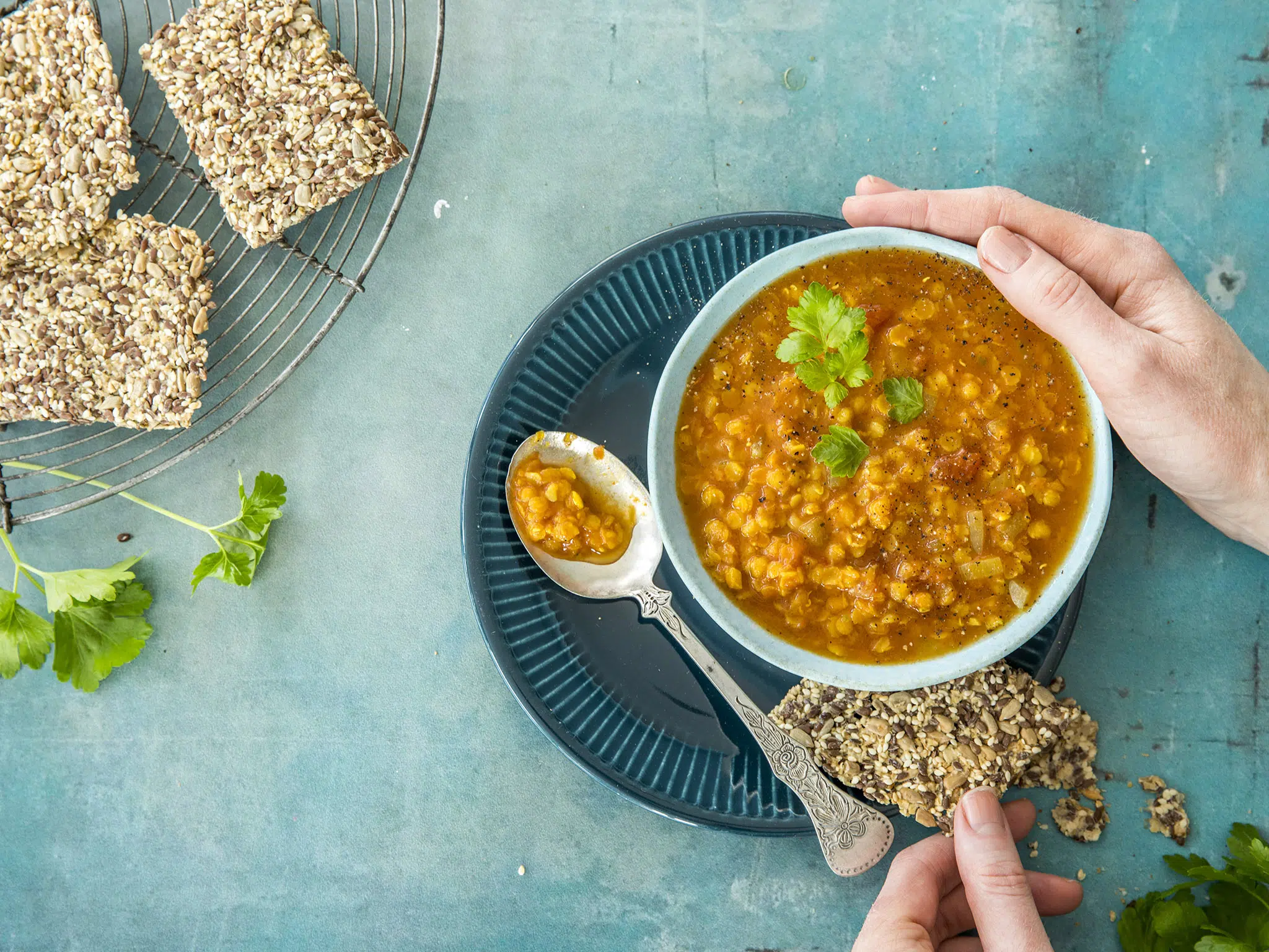 En person holder en kjeks med frø og en skål med oransje linsesuppe pyntet med persille, med flere kjeks og persille på en blå flate.