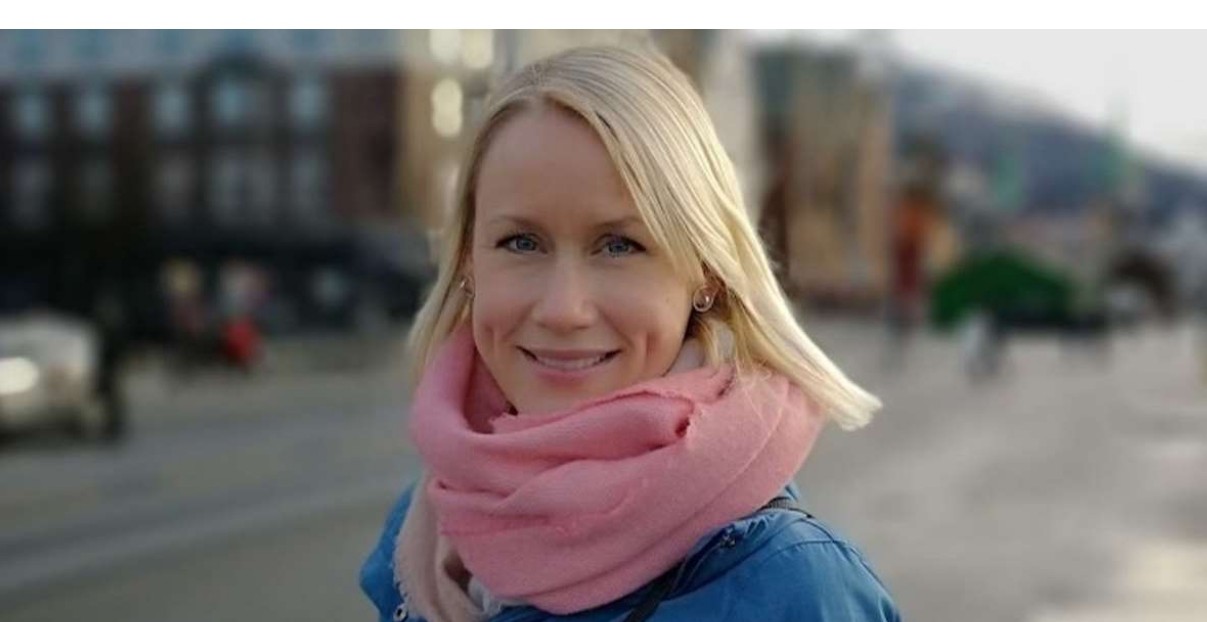 En kvinne med blondt hår, iført blå jakke og rosa skjerf, står utendørs i en bygate med uskarpe bygninger i bakgrunnen.