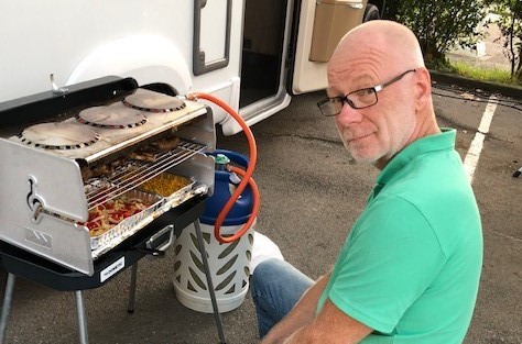 En mann med briller og grønn skjorte sitter ved en liten utegrill med mat på brett ved siden av en parkert bobil.