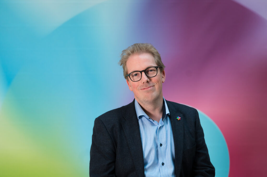 En mann med briller, blå skjorte og mørk blazer står foran en fargerik bakgrunn med gradienter.