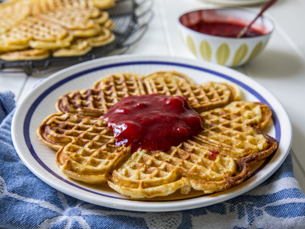 En tallerken med en blomsterformet vaffel toppet med jordbærsaus, med flere vafler på et kjølerist og en bolle med saus i bakgrunnen.