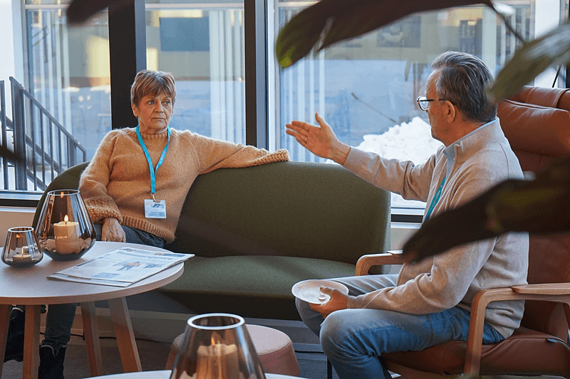 To eldre voksne sitter i en salong og snakker sammen, den ene gestikulerer med hånden mens den andre lytter. På bordet foran dem står det stearinlys og en avis.