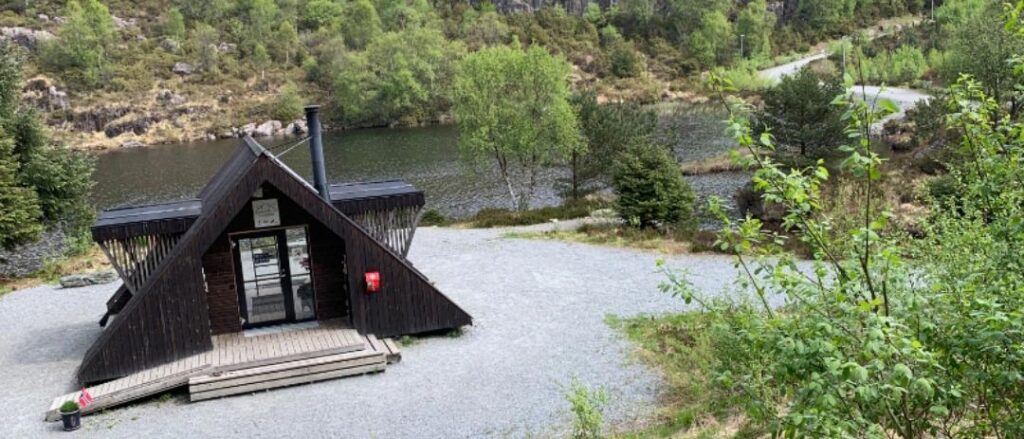 En liten, mørk trebygning i A-form med skorstein ligger ved siden av en grussti nær en elv, omgitt av trær og grøntområder.
