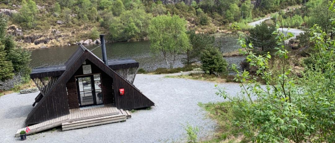 En liten, mørk trebygning i A-form med skorstein ligger ved siden av en grussti nær en elv, omgitt av trær og grøntområder.