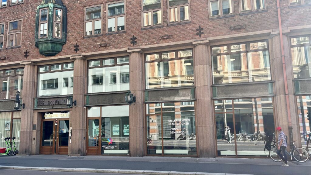 Gatebilde av en murbygning med store vinduer, steinsøyler, en mann på sykkel og refleksjoner av en annen bygning i glasset.