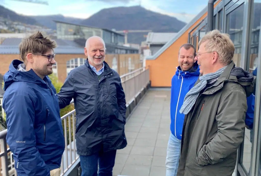 Fire menn i jakker står og snakker sammen på en utendørs balkong med bygninger og et fjell synlig i bakgrunnen.