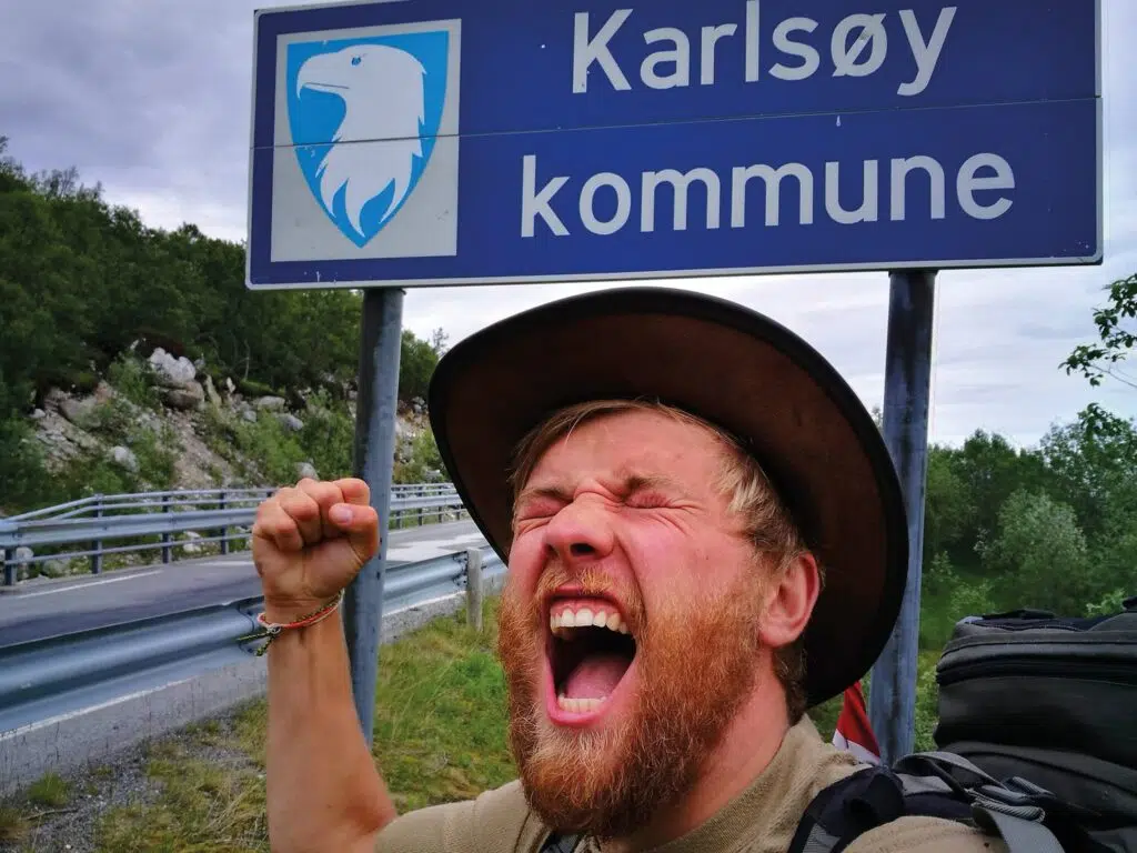 En mann med skjegg og hatt roper opphisset og hever neven foran et skilt med påskriften "Karlsøy kommune" ved siden av et blått ørneemblem.
