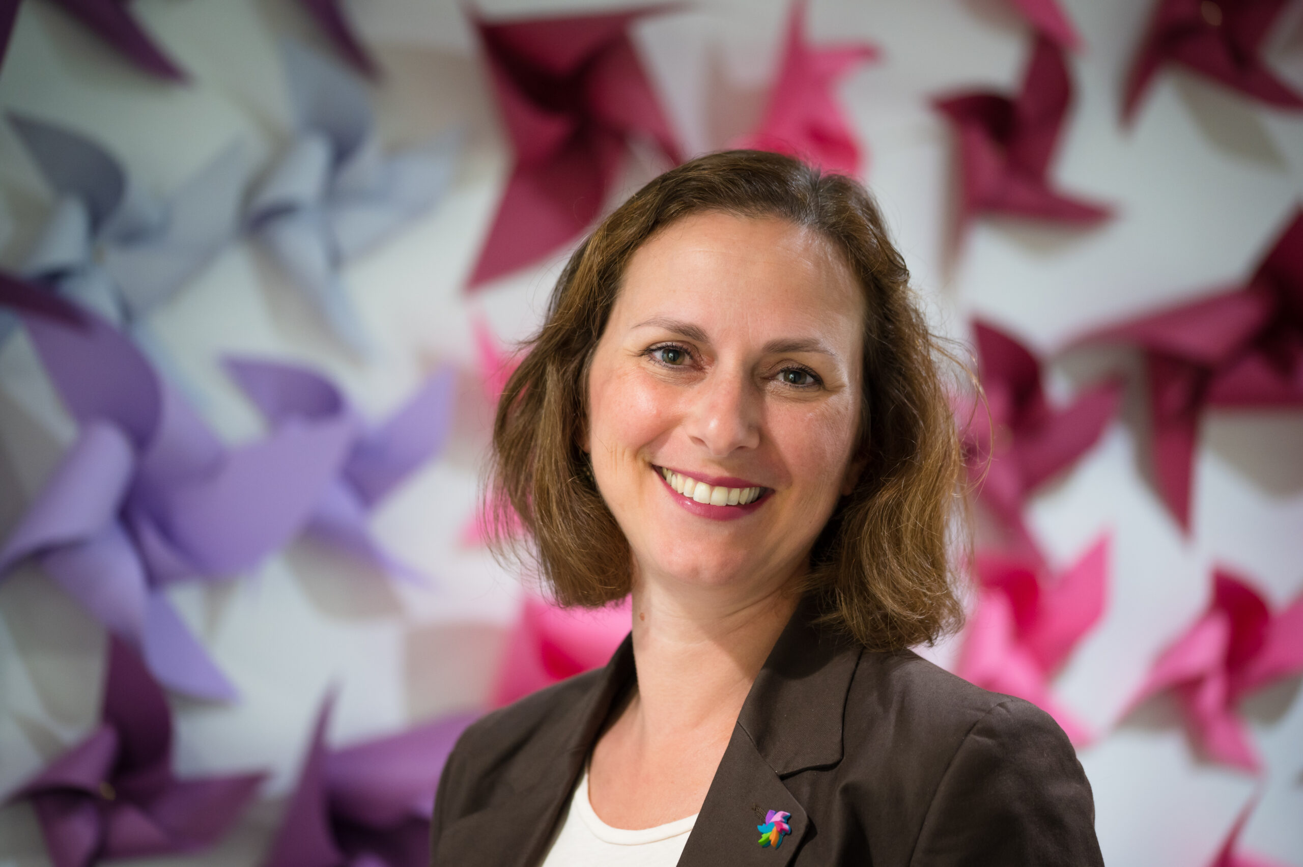 En kvinne med brunt hår smiler til kameraet, stående foran en vegg dekorert med rosa og lilla papirhjul.