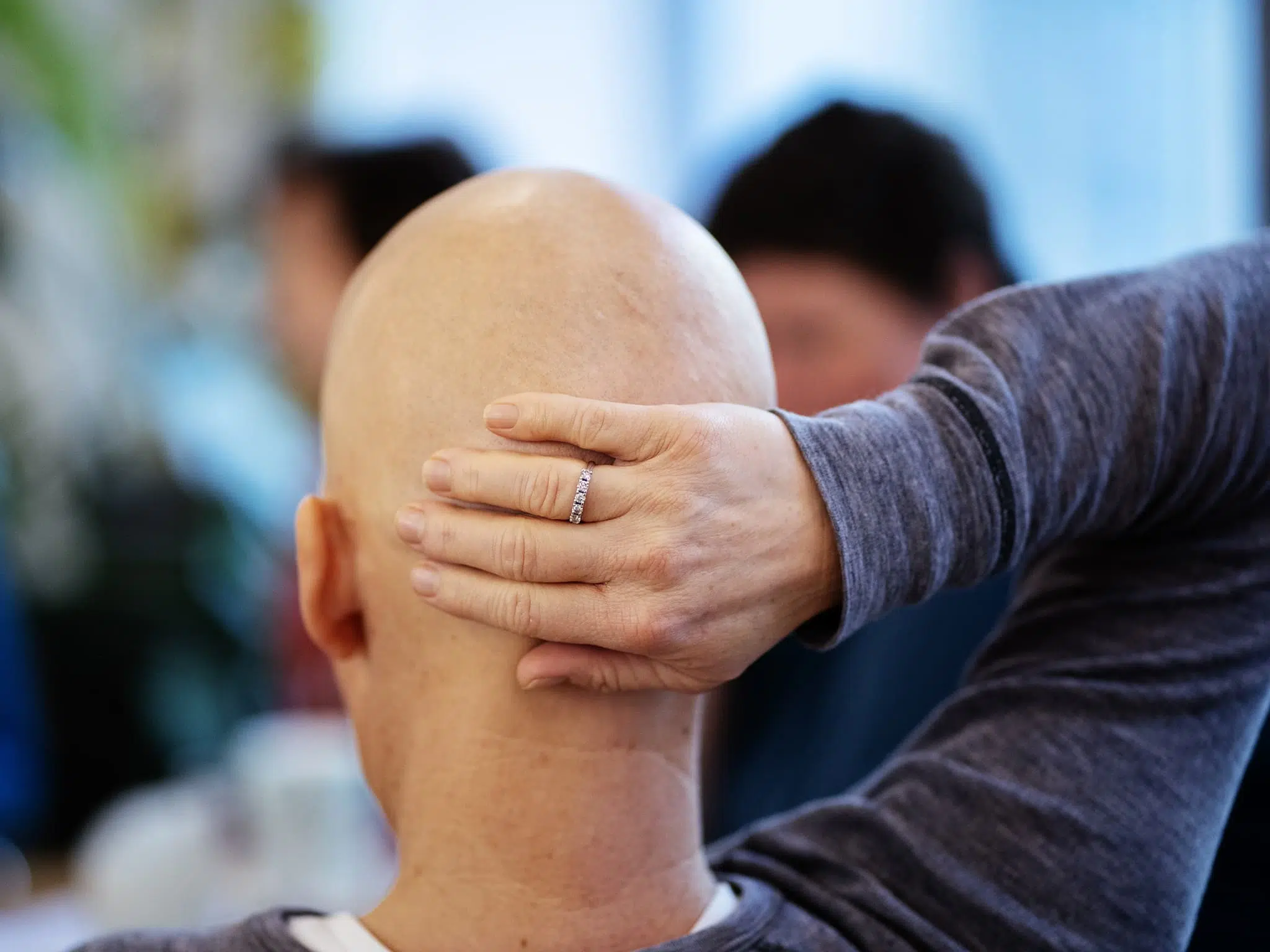 En person med skallet hode sitter med ryggen til kameraet og hviler hånden på bakhodet. En ring er synlig på fingeren.