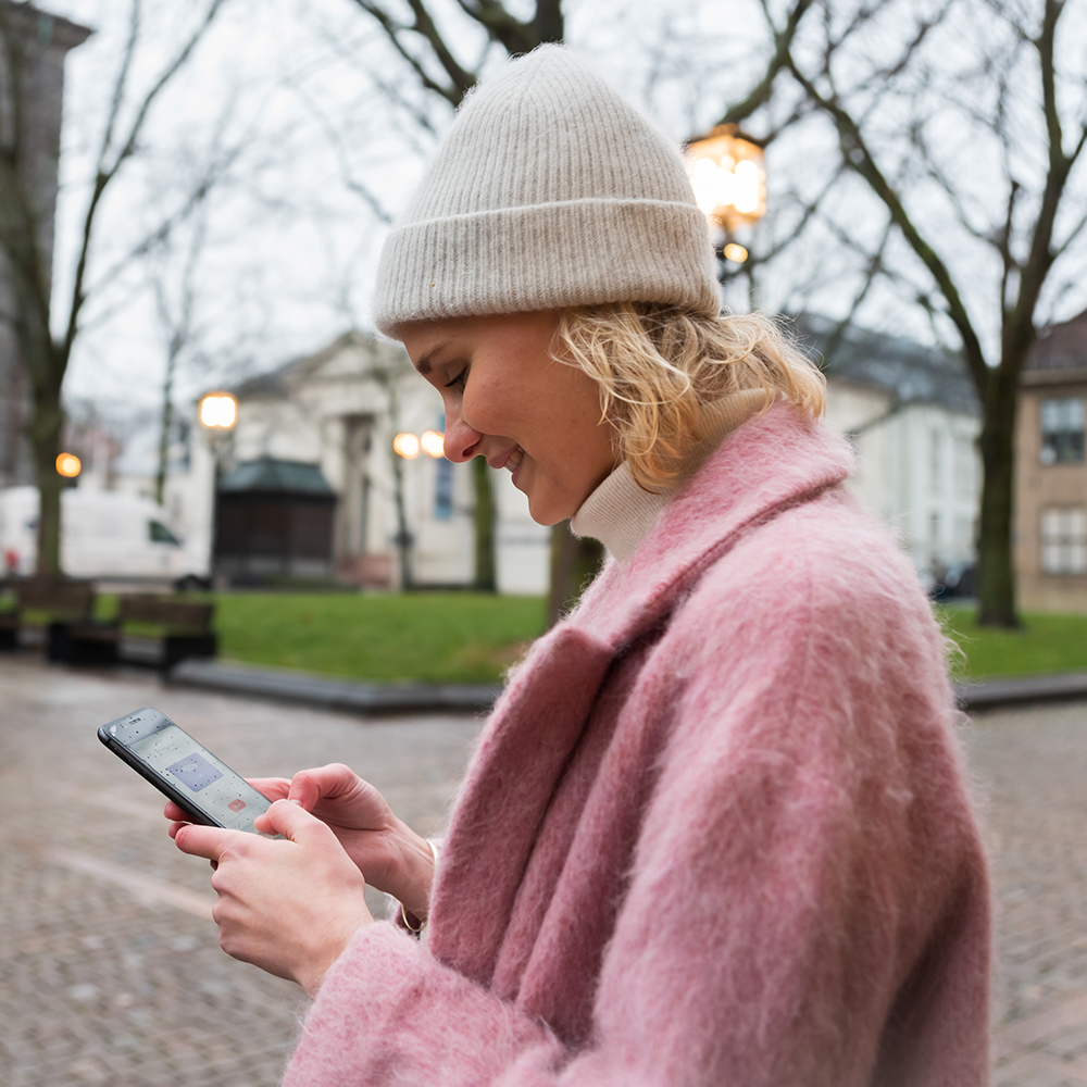En person med lys strikkelue og rosa kåpe står utendørs og smiler mens hun bruker smarttelefonen på en overskyet dag.