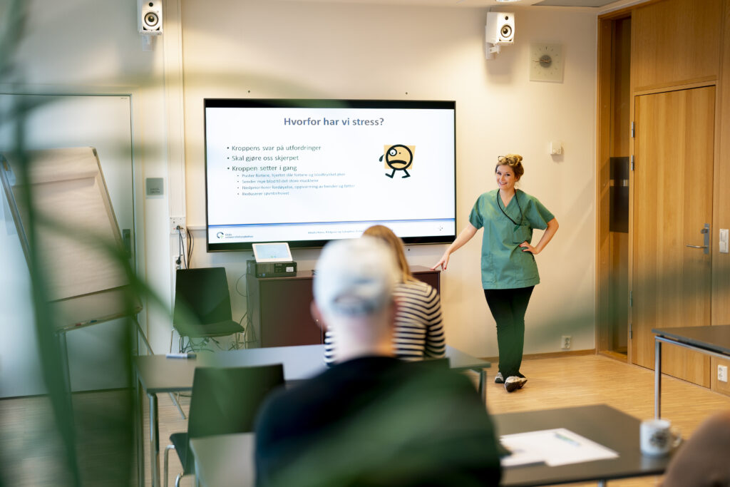 En kvinne i operasjonsklær står ved siden av en presentasjonsskjerm i et klasserom og holder et foredrag om stress for de sittende deltakerne.