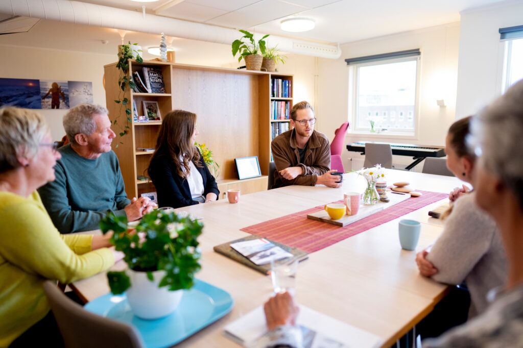 Seks personer sitter rundt et bord i et lyst rom og snakker sammen. På bordet står kopper, planter og papirer, og i bakgrunnen er det hyller og vinduer.