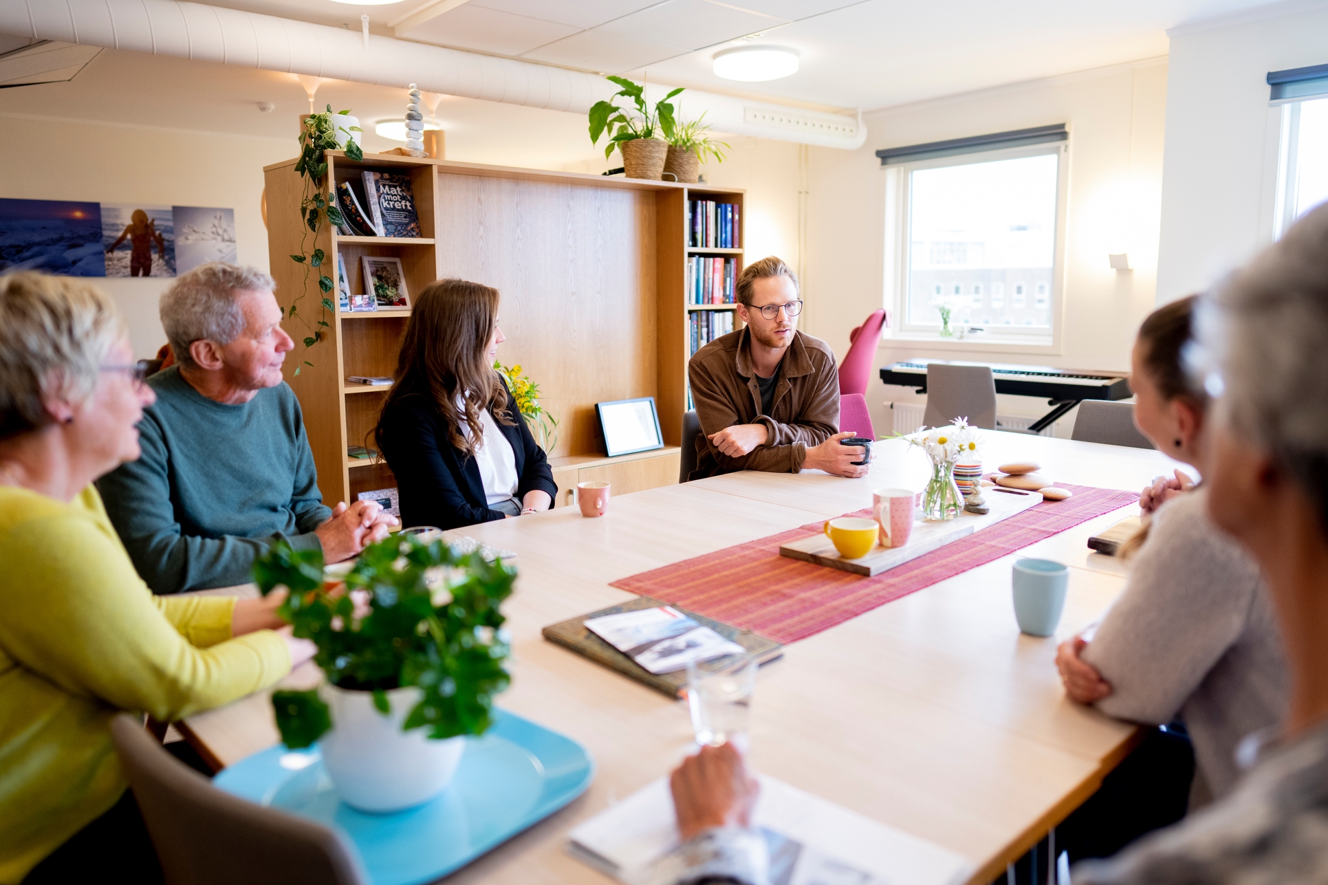 Seks personer sitter rundt et bord i et lyst rom og snakker sammen. På bordet står kopper, planter og papirer, og i bakgrunnen er det hyller og vinduer.