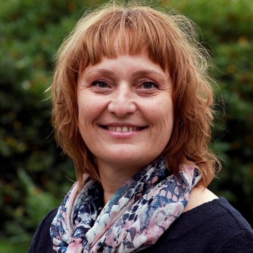 En kvinne med kort, bølget, rødbrunt hår smiler til kameraet, iført mønstret skjerf og mørk overdel, med grønne planter i bakgrunnen.