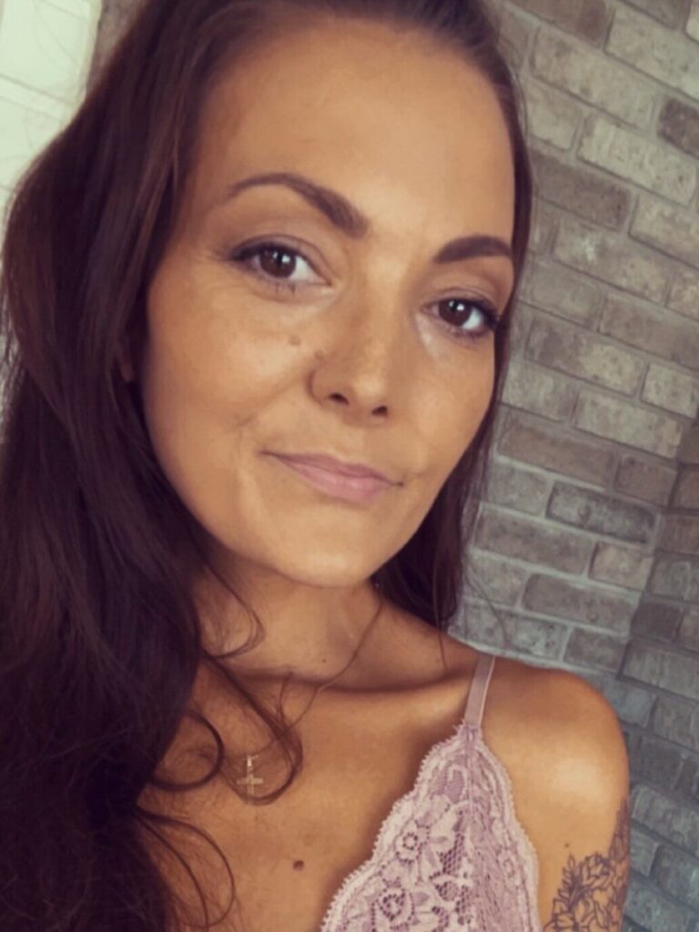 En kvinne med langt brunt hår og blondetopp poserer foran en murvegg, mens hun ser nøytralt inn i kameraet.