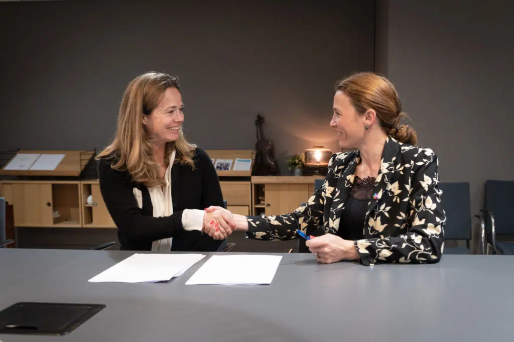 To kvinner sitter ved et bord, håndhilser og smiler, med dokumenter og en penn foran seg, noe som tyder på en forretningsavtale eller kontraktsignering.