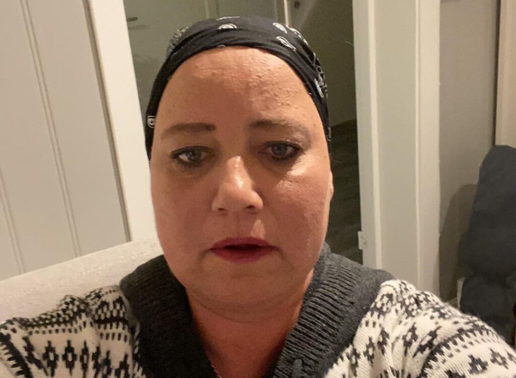 En kvinne med svart skaut og mønstret genser ser inn i kameraet innendørs, med en hvit vegg og en åpen dør i bakgrunnen.