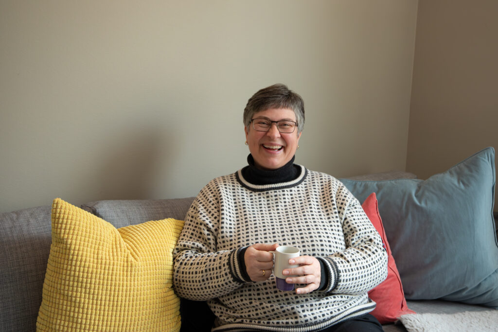 En person med kort hår og briller sitter i en grå sofa, smiler og holder i et krus. Ved siden av dem ligger gule, røde og blå puter.