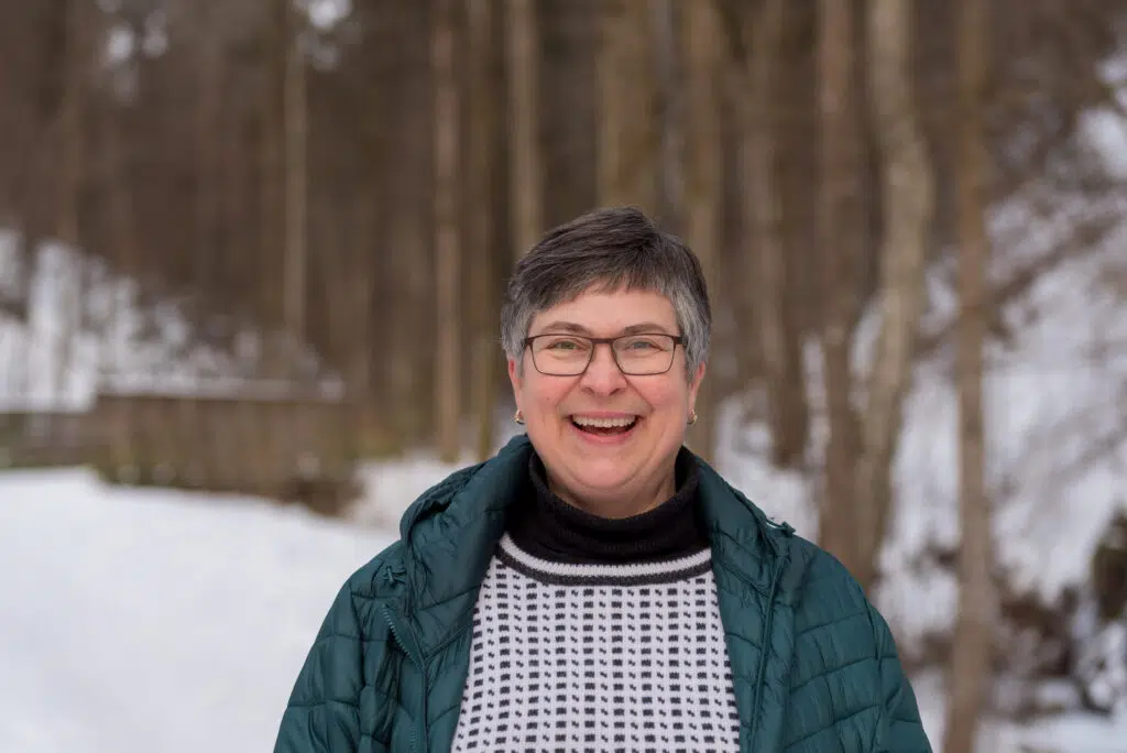 En person med kort, grått hår og briller smiler utendørs, iført grønn jakke og mønstret genser, med en snødekt skog i bakgrunnen.