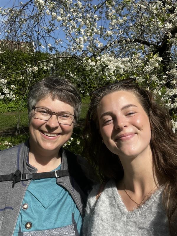 To kvinner smiler utendørs i sterkt sollys, stående foran et blomstrende tre med hvite blomster.