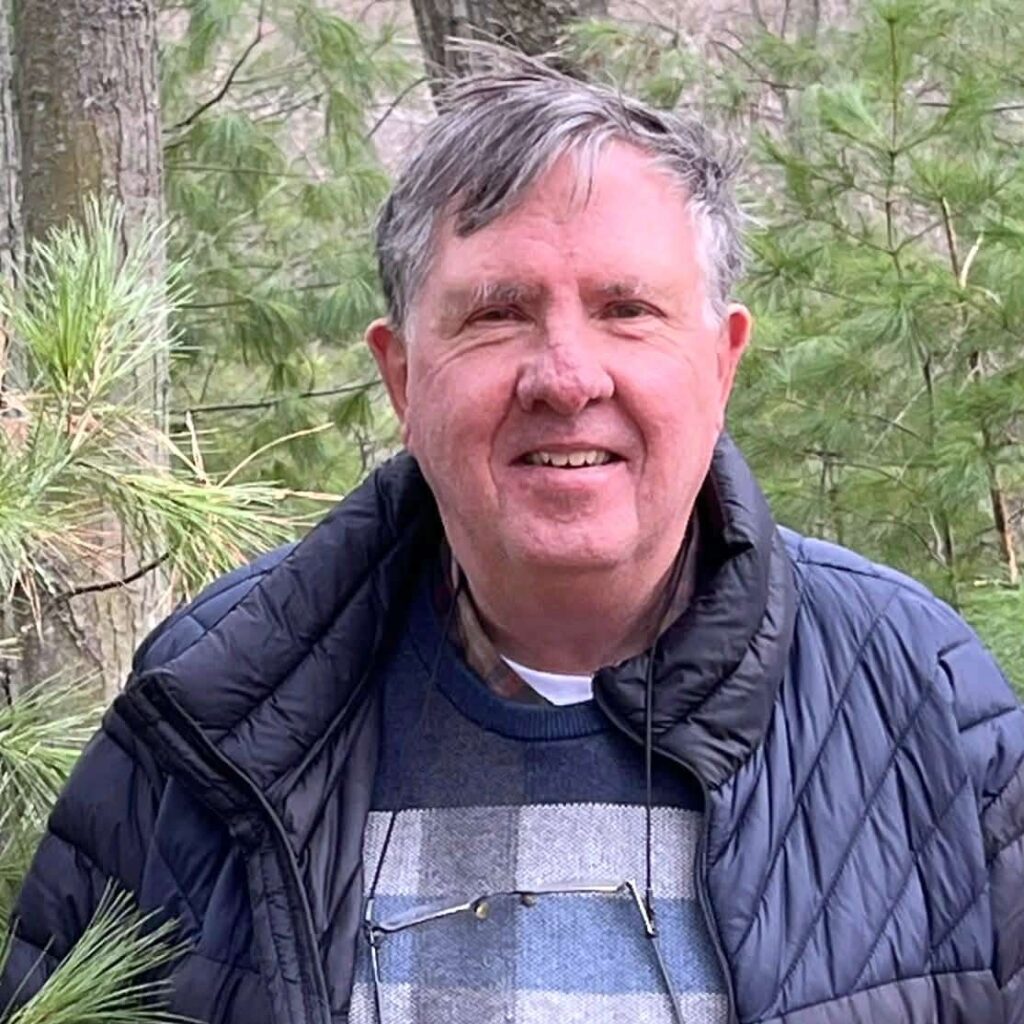 En eldre mann med grått hår står utendørs foran grønne furutrær, iført mørk jakke og mønstret genser.