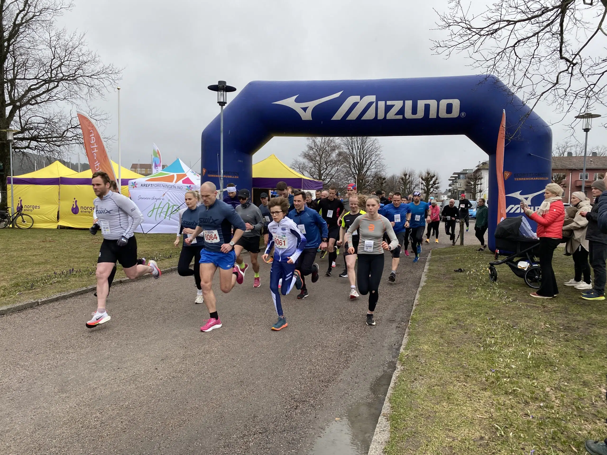 En gruppe løpere starter et løp under en blå oppblåsbar Mizuno-bue, med tilskuere og arrangementstelt synlige i bakgrunnen på en overskyet dag.